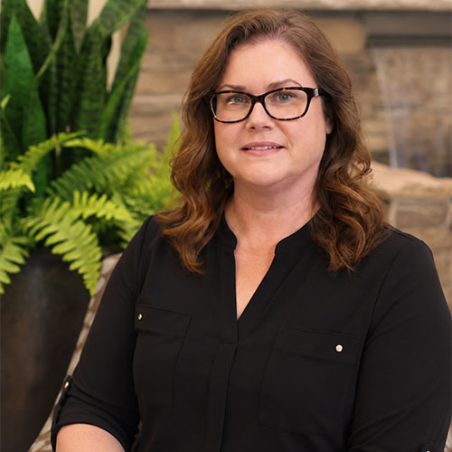 Woman with glasses and shoulder-length brown hair, wearing a V-neck business top.