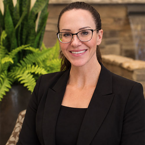 Smiling woman wearing glasses, a round neck top and jacket with her long dark hair pulled back in a ponytail.