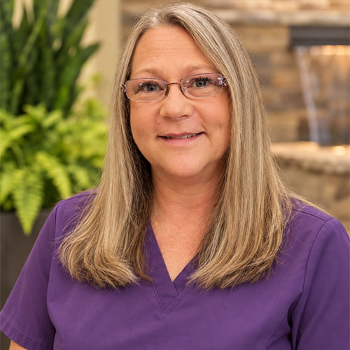 Smiling woman with straight, long, blonde hair wearing glasses and a V-neck scrub top.